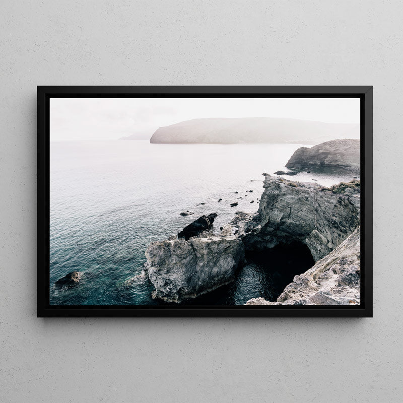 A framed landscape photograph of a rocky coastal cave and sea cliffs, displayed on a light gray textured wall. The print features a black frame and showcases a scene with deep blue water, gray rocks, and a hazy distant island.