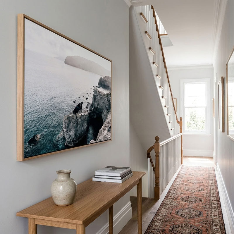 A large framed landscape photograph of a rocky coastline hanging on a light gray wall in a bright hallway with a wooden console table, ceramic vase, and staircase.