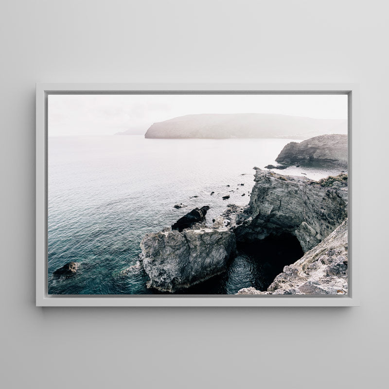 A framed photographic print of a misty coastal landscape with dark rocky cliffs, a sea cave, and calm blue-gray water, displayed against a light gray wall.