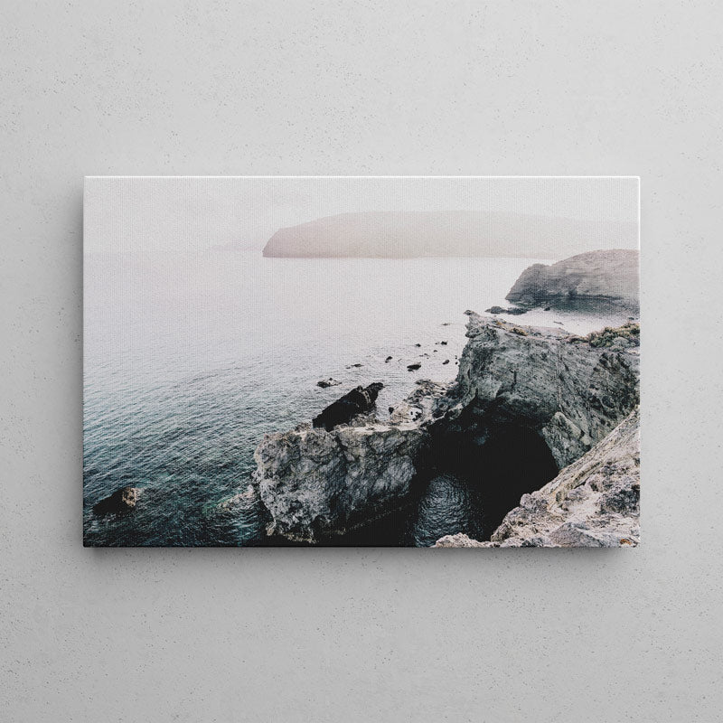 A rectangular wall art print showing a rocky coastal cave and sea scene, mounted against a light gray textured wall.