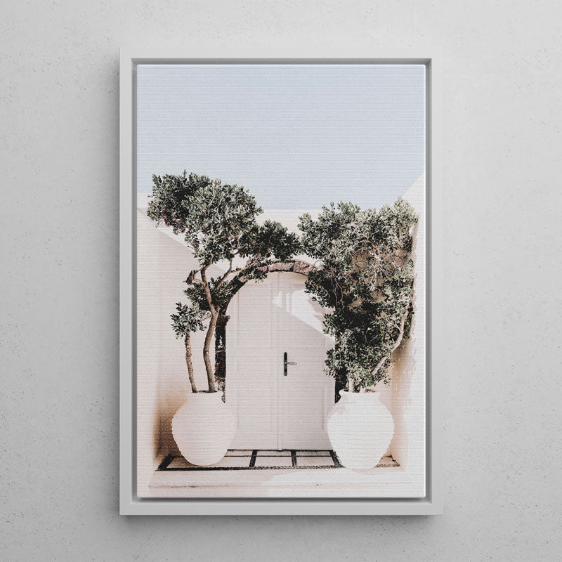 A framed art print featuring a Mediterranean courtyard scene with a white door, olive trees, and white planters, mounted on a light gray textured wall.