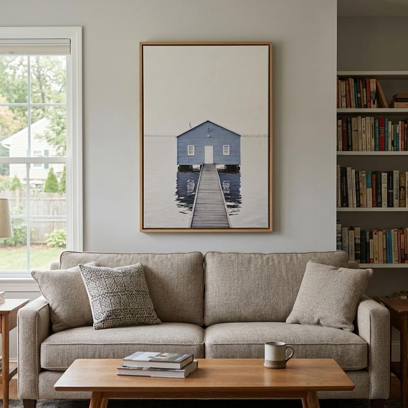 A framed art print of a blue wooden dockhouse on a pier, displayed on a light gray wall above a beige fabric sofa in a living room setting.