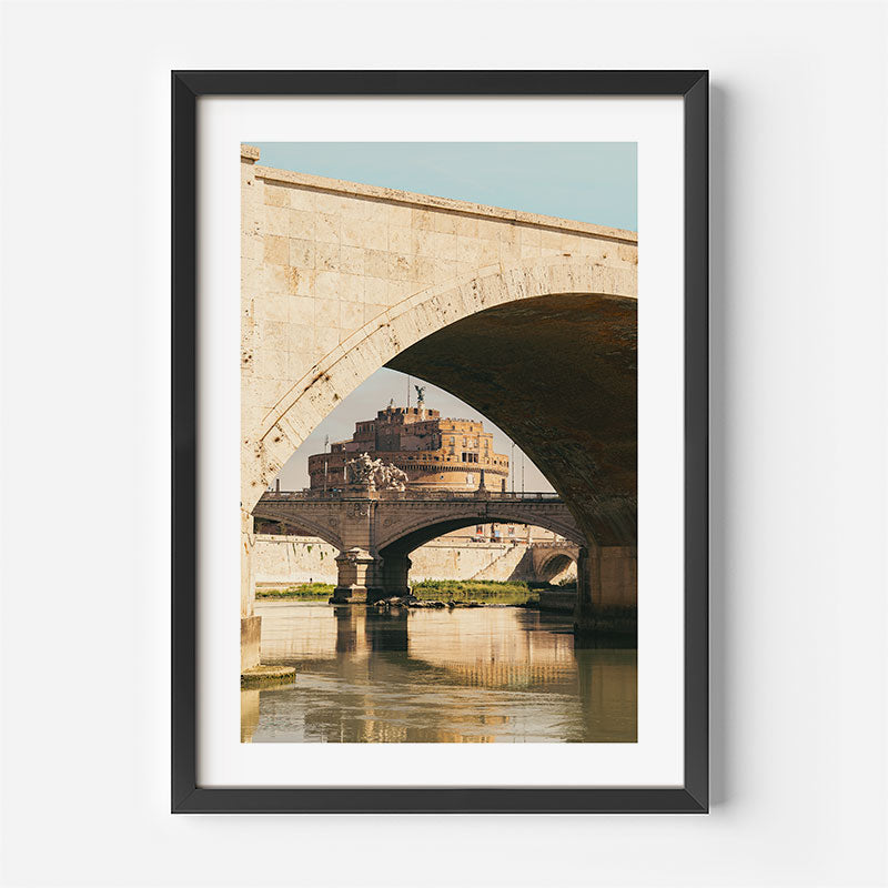 A framed art print featuring a scenic photograph of Castel Sant'Angelo in Rome, viewed through the arch of a stone bridge over the Tiber River. The print is framed in a simple black frame with a white mat, set against a clean white background.