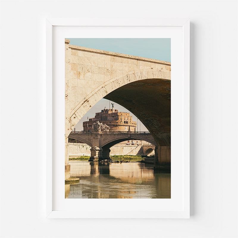 A framed art print depicting the historic Castel Sant'Angelo in Rome under a stone arch bridge, with reflections in the water, presented in a white frame against a light gray wall.