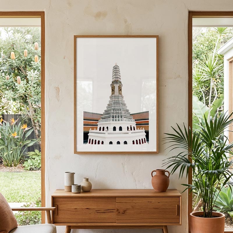 A framed art print of a Thai temple tower hanging on a textured beige wall above a wooden console table, flanked by two windows with outdoor greenery and a potted palm.