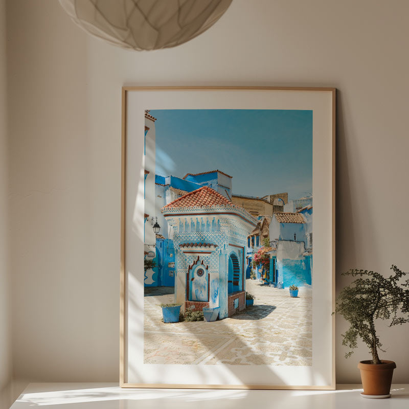 Framed artwork of a colorful street scene on a wall with a plant below.