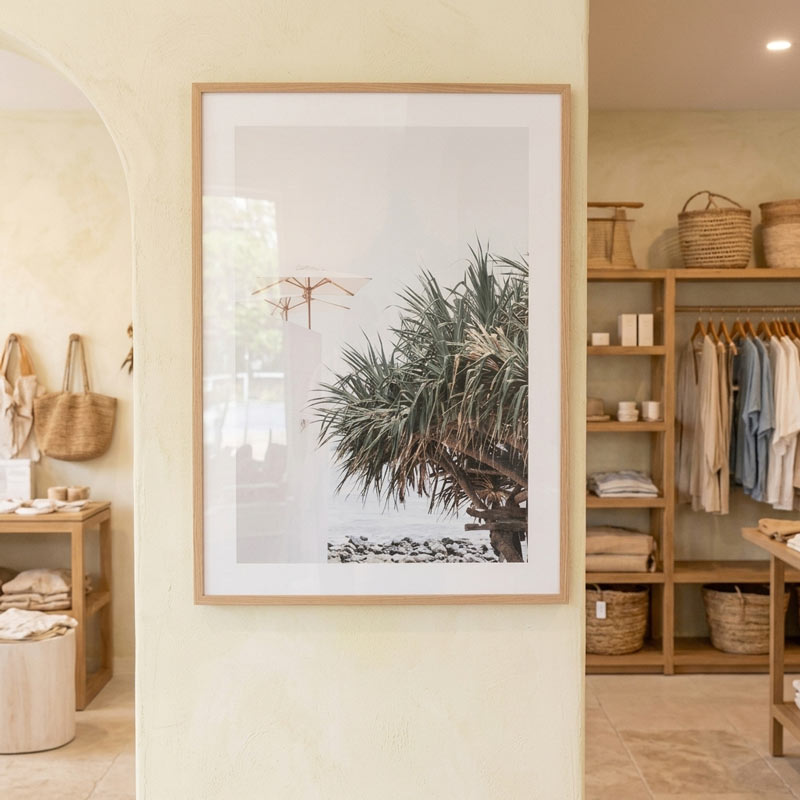 A framed botanical art print featuring a palm tree and a small dragonfly, displayed on a beige wall inside a boutique interior.