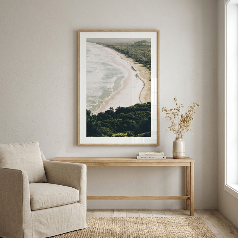 A framed vertical art print of an aerial beach landscape hangs on a light gray wall above a wooden console table in a styled living room setting with a beige armchair and a vase.