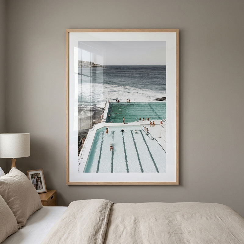 A framed art print depicting an ocean pool scene, styled in a bedroom setting above a bed with neutral-colored bedding and a bedside lamp.