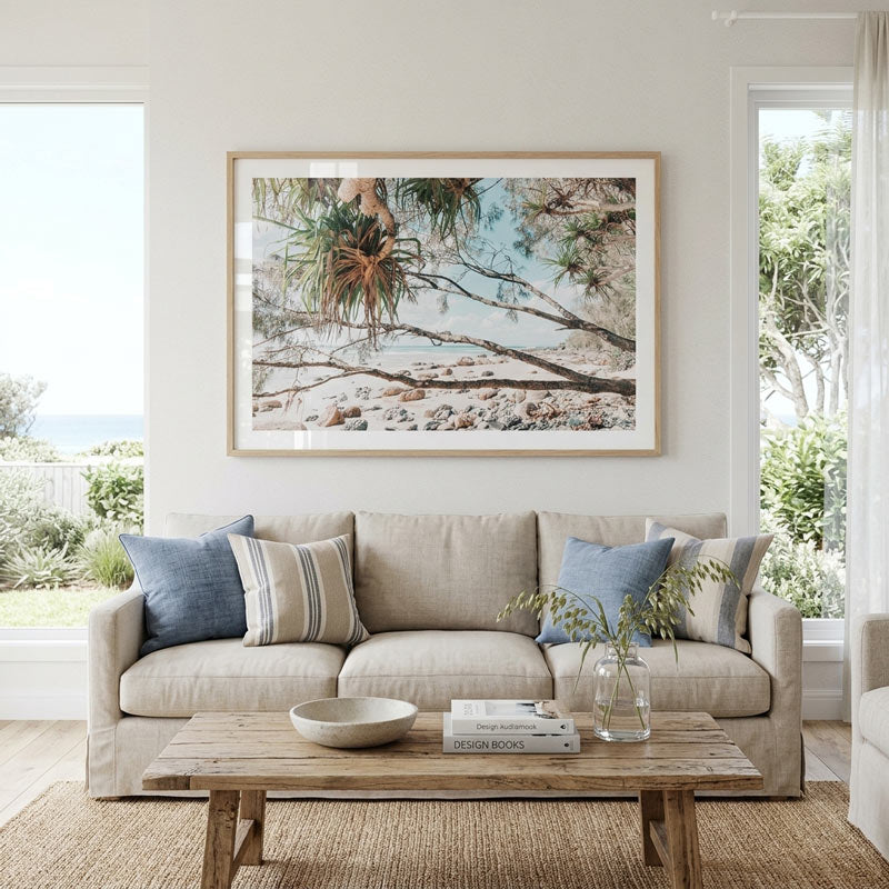 A large framed beach and plant print hangs above a beige sofa in a bright, modern living room with blue and striped accent pillows, a wooden coffee table, and windows looking out to greenery.