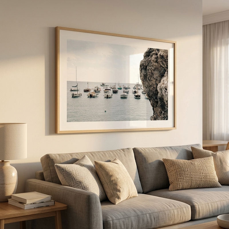 A large framed landscape photograph of a coastal harbor with sailboats and a rocky cliff, displayed on a beige wall above a gray sofa in a modern living room setting.