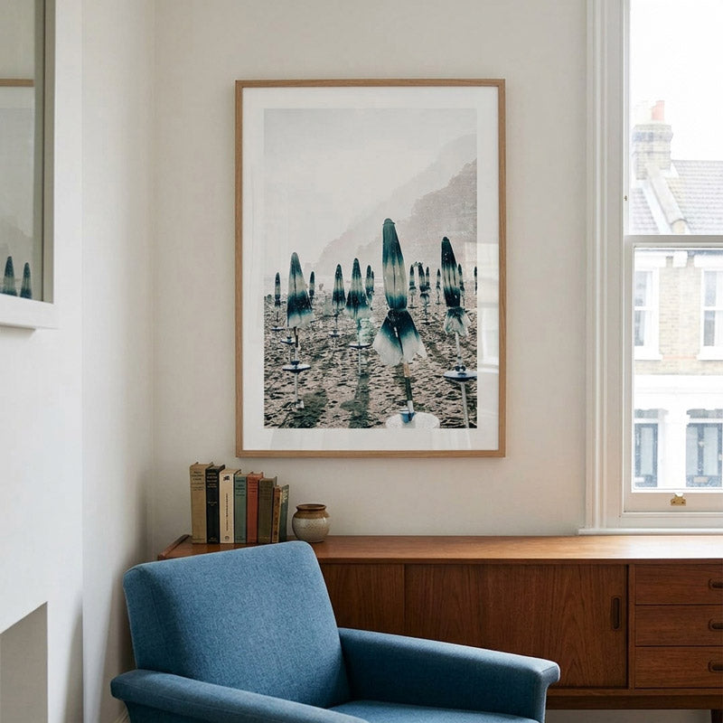 A framed art print featuring a moody, misty landscape with blue and gray rock spires is displayed on a light beige wall above a mid-century modern wooden credenza and a blue armchair.