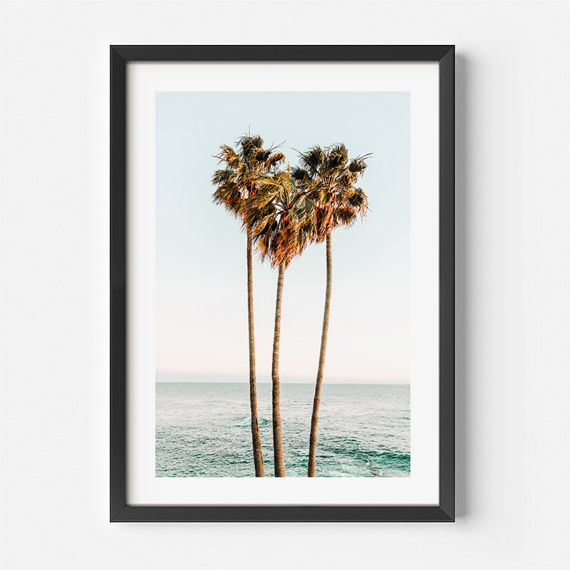 Framed artwork of three palm trees by the ocean on a white wall.