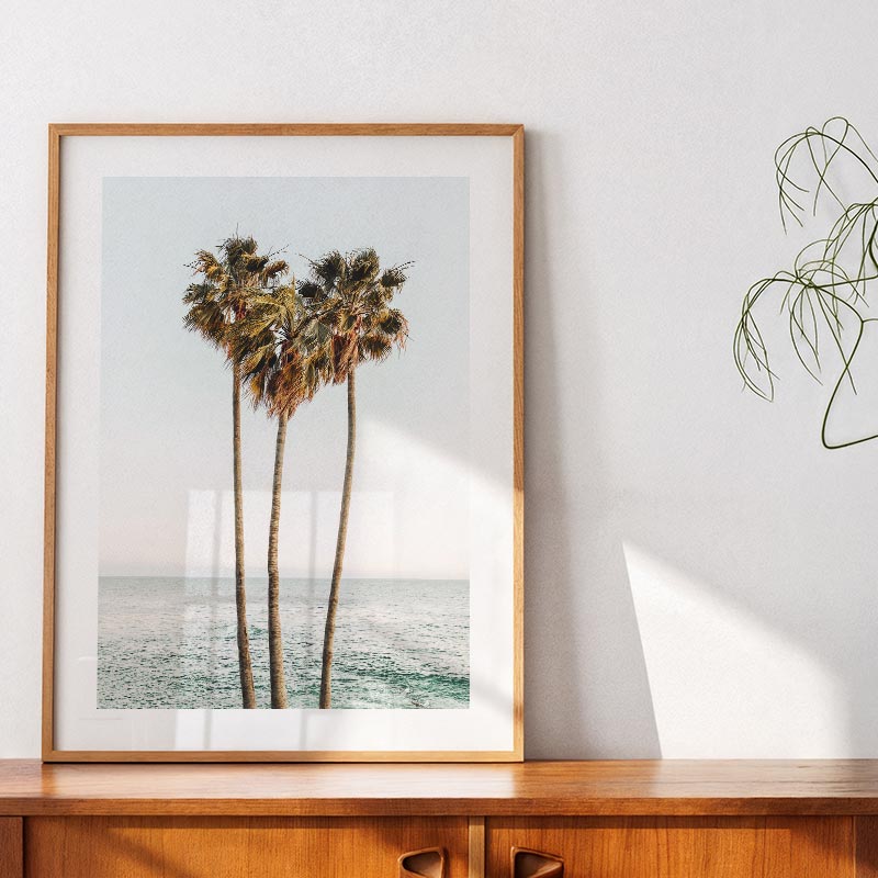 Framed artwork of palm trees by the ocean on a white wall.