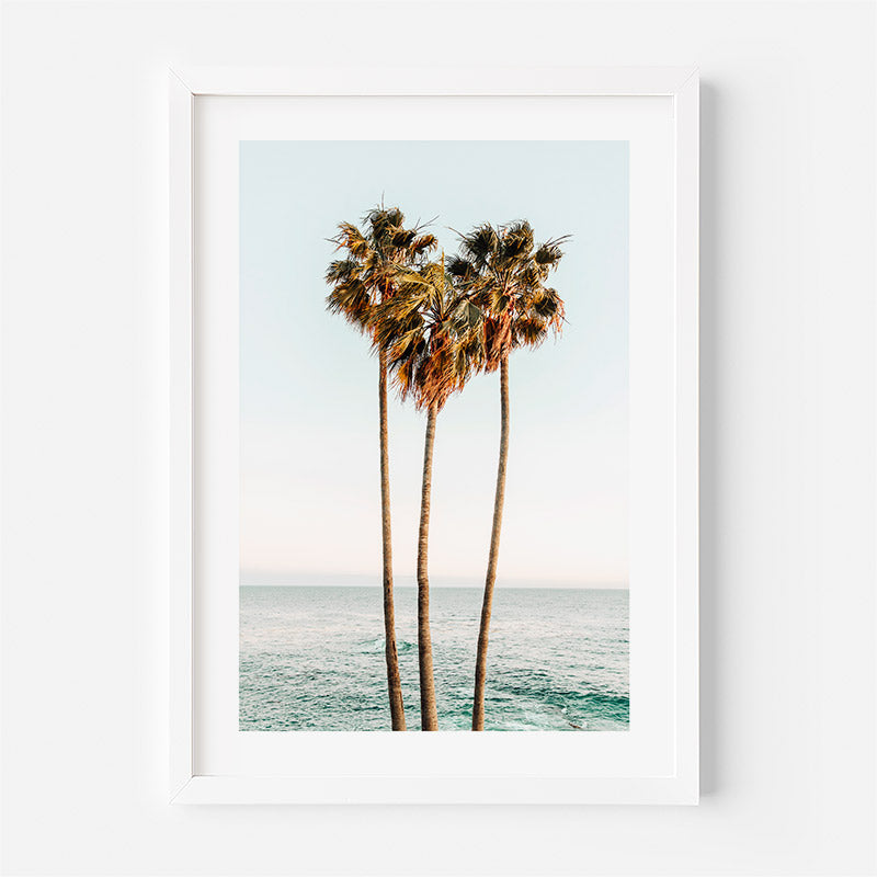 Framed artwork of three palm trees by the ocean on a white wall.