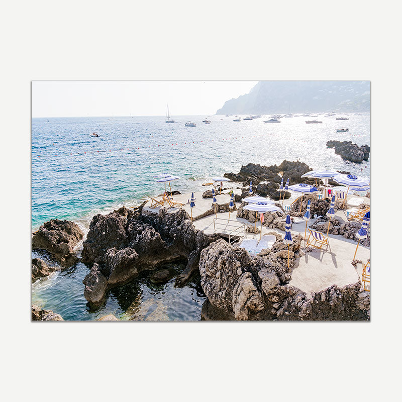 Beach scene with rocky shoreline and people under umbrellas, with boats in the distance.