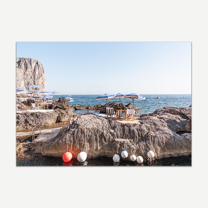 Beach scene with lounge chairs on rocks, umbrellas, and floating buoys in the water.