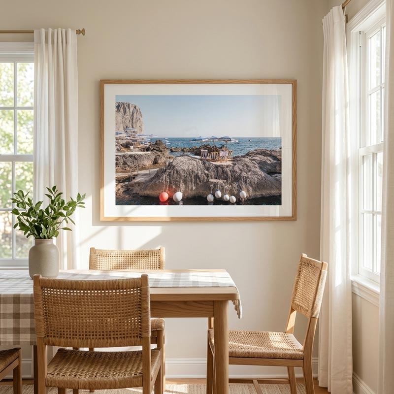 A framed coastal landscape photograph hangs on a beige wall above a wooden dining table and woven chairs in a bright, sunlit room.