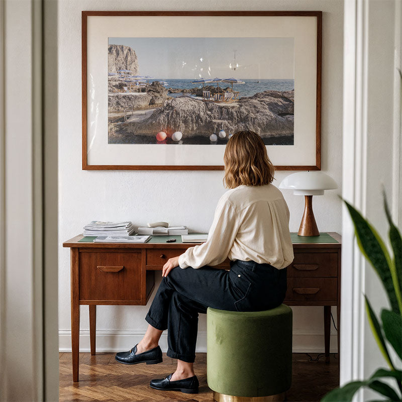 Woman sitting on a stool at a desk in a room with a large framed picture on the wall.