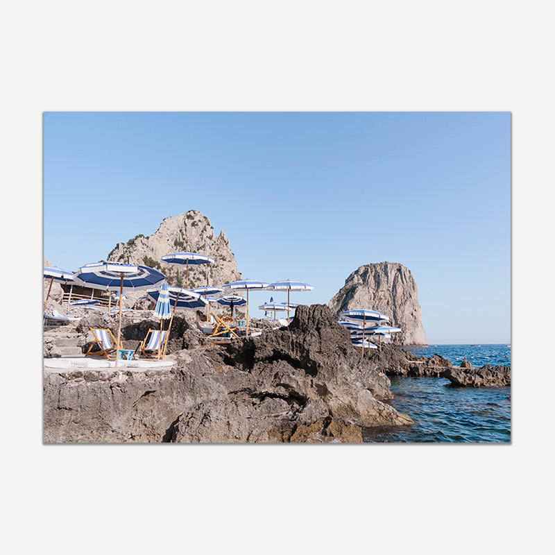 Beach chairs and umbrellas on a rocky cliff overlooking the ocean with clear blue sky.