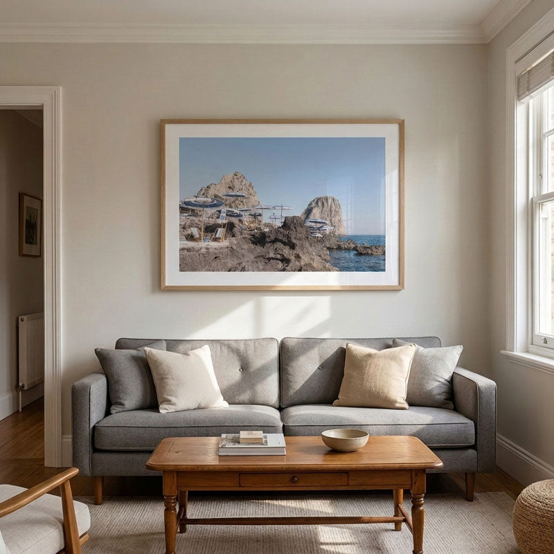 A framed coastal landscape print hangs on a light beige wall above a gray sofa in a bright, modern living room with a wooden coffee table and neutral decor.