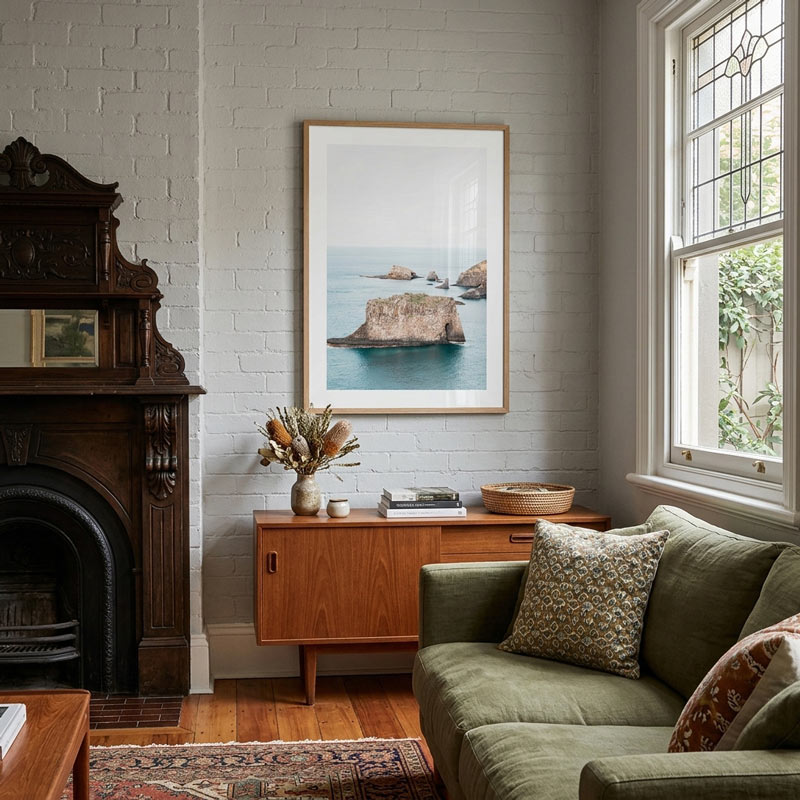 A framed art print depicting sea stacks in a coastal bay hangs on a white brick wall in a styled living room with an olive green sofa, a mid-century wooden sideboard, and a dark wood fireplace.