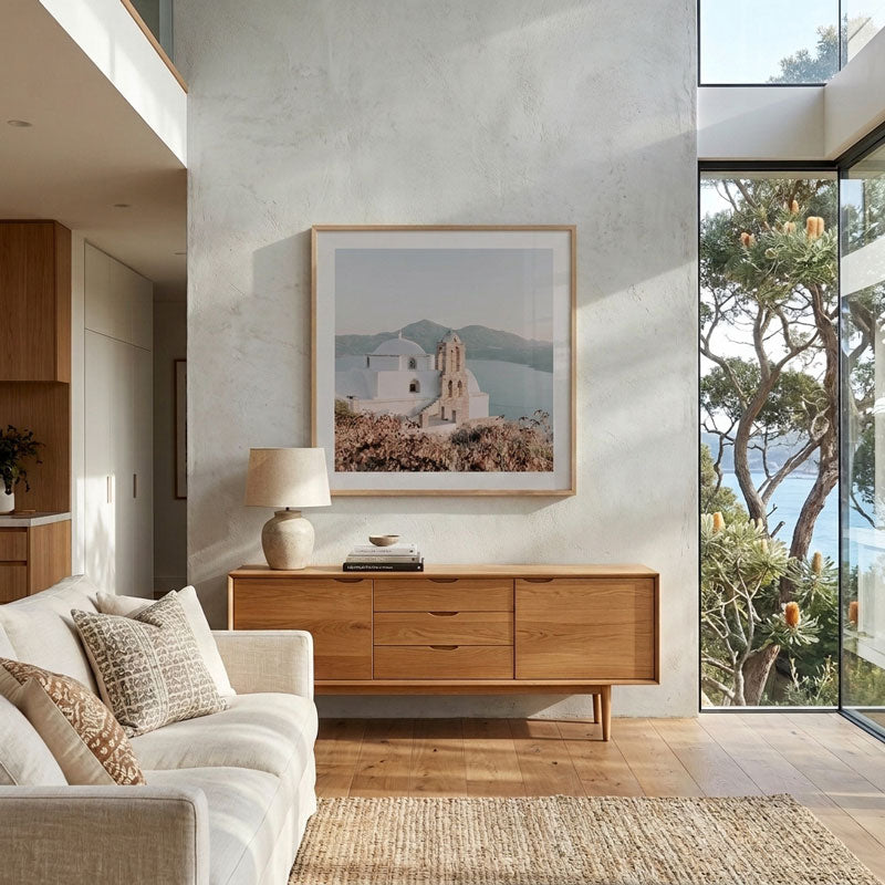 A framed art print depicting a white church on a hillside with blue water and mountains, displayed on a textured light gray wall in a modern, sunlit living room with a wooden sideboard and a white sofa.