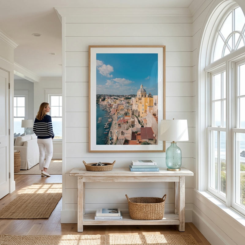 A large framed art print depicting a coastal city with blue water and warm-toned buildings, displayed on a white shiplap wall above a console table in a bright, modern room.