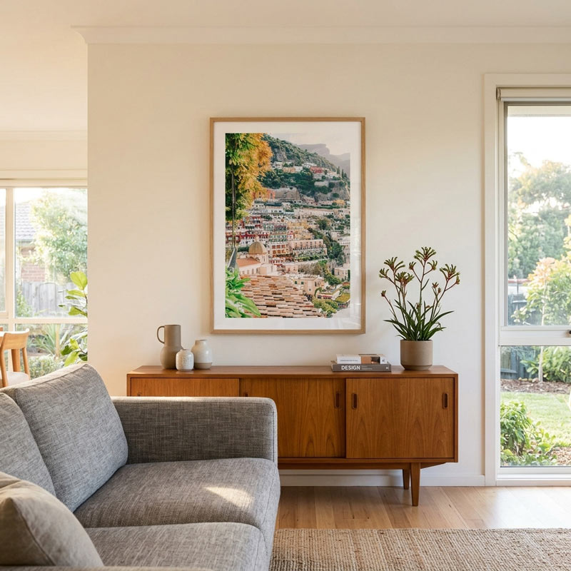 A framed art print depicting a scenic hillside village with colorful houses and greenery, displayed on a cream wall above a mid-century modern wooden sideboard in a bright, modern living room.