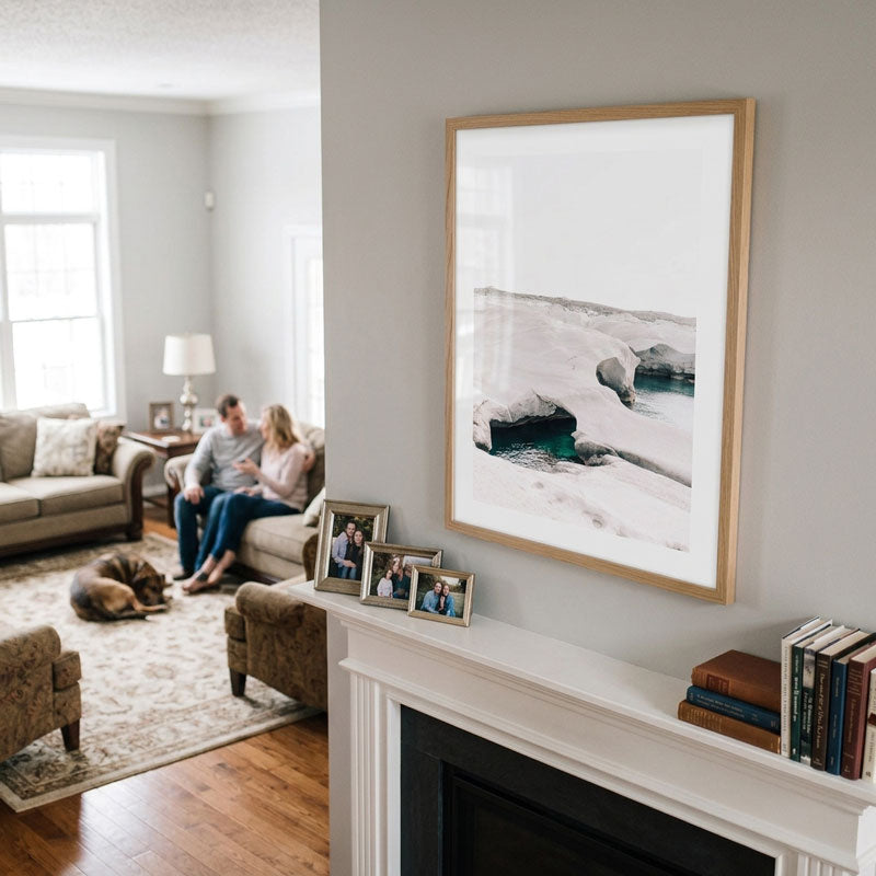 A large framed landscape art print with a light wood frame hangs on a white wall above a fireplace mantel. The print depicts a snowy icy shoreline with dark rocks.