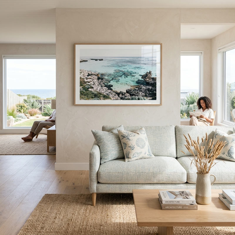 A framed coastal landscape print hangs on a beige wall above a light gray sofa in a bright, modern living room. The artwork shows rocky shoreline and turquoise water.