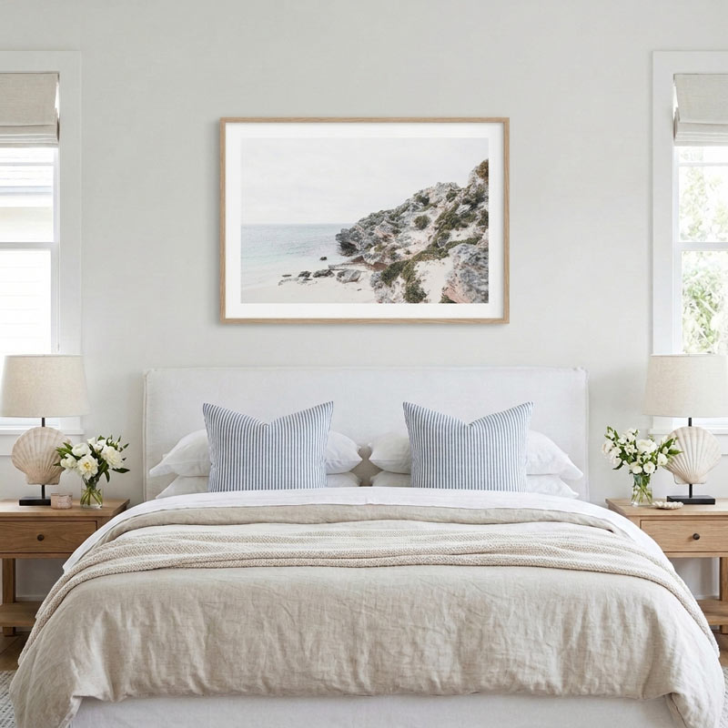 A framed art print of a coastal rocky cliff and sea scene, displayed on a light gray wall above a neatly made bed with beige linens and blue striped pillows in a bright, modern bedroom.