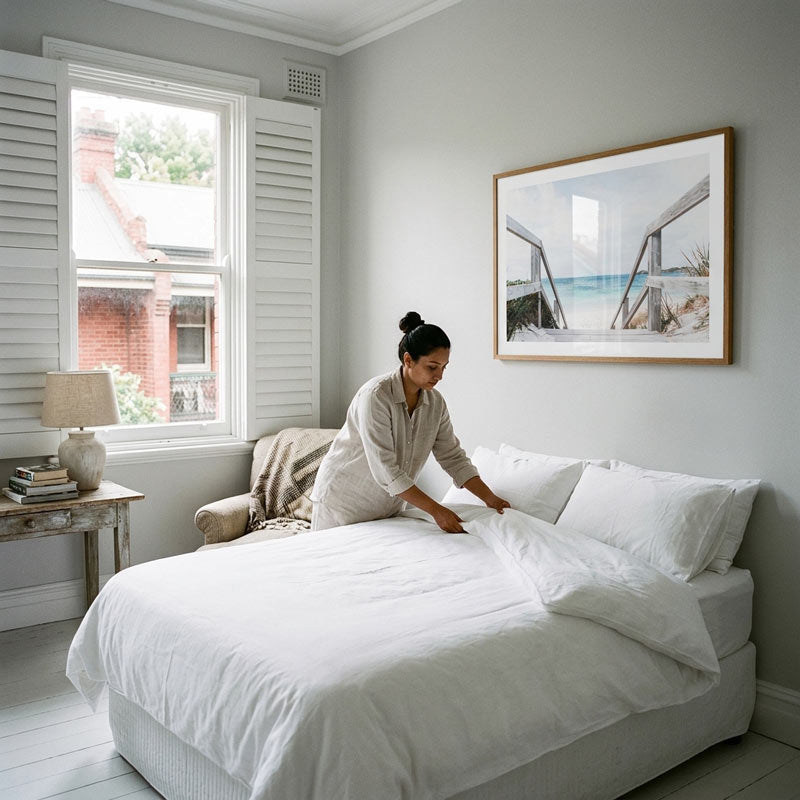A woman in a light-colored robe is making the bed with white linens in a bright, modern bedroom with white shutters and a framed print on the wall.