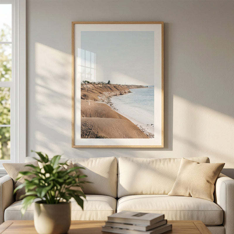 A framed coastal landscape art print hangs on a light gray wall above a cream sofa in a sunlit living room. The print shows sandy dunes, a beach, and blue water, with a light wood frame and white mat.
