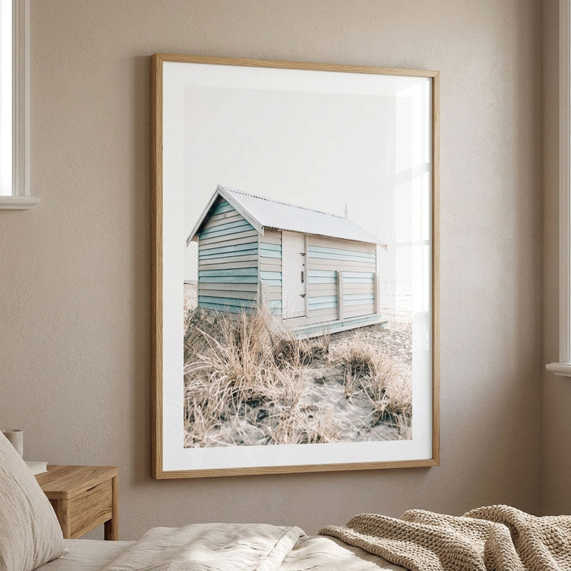 A framed beach art print hanging on a beige wall above a bed. The print shows a small weathered blue beach hut surrounded by dry grass, enclosed in a light wood frame with a white mat.