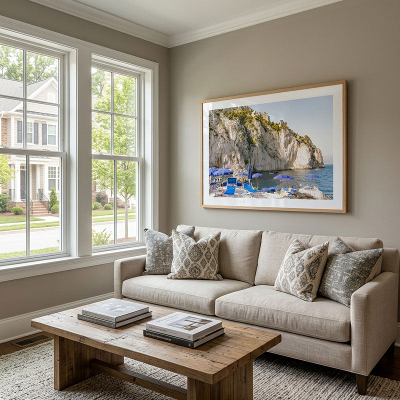A framed coastal landscape print of a rocky beach and blue umbrellas hangs on a beige wall above a light beige sofa in a bright, modern living room with a wooden coffee table and large windows.