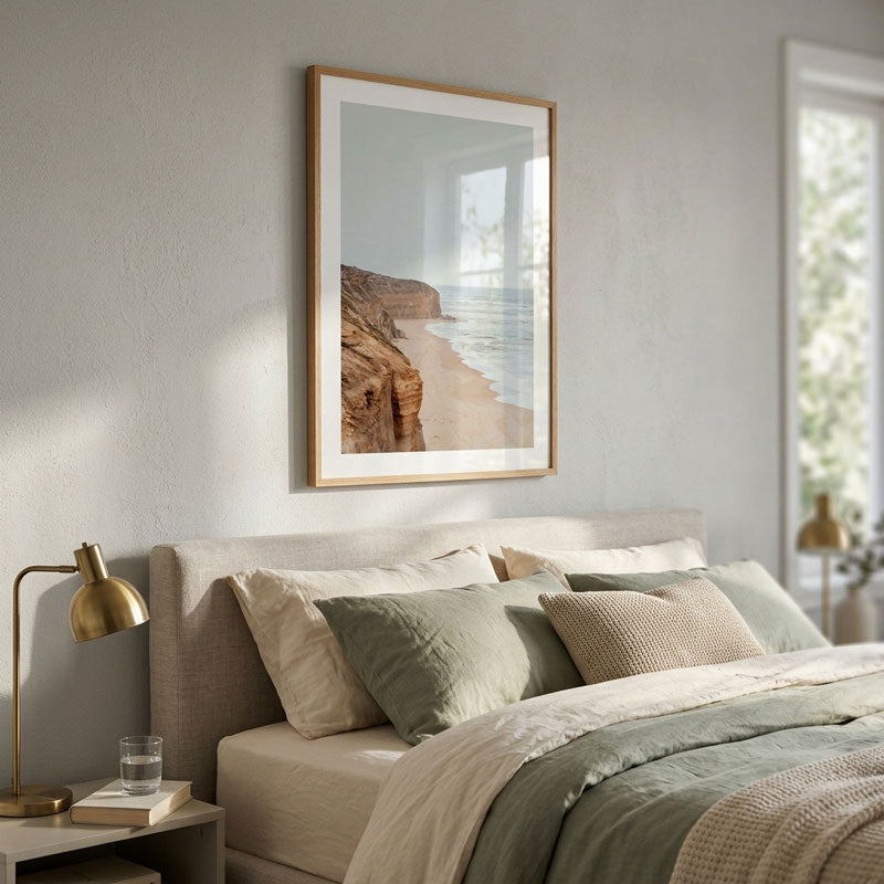 A framed vertical art print depicting a coastal beach and rocky cliffs hangs on a textured light gray wall above a neatly made bed with sage green and beige bedding in a modern, softly lit bedroom.