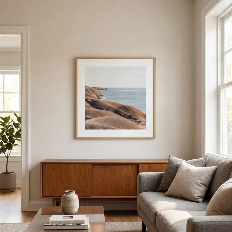 A framed landscape art print of coastal hills and the sea hangs on a beige wall in a modern living room setting above a mid-century wooden sideboard and a gray sofa.