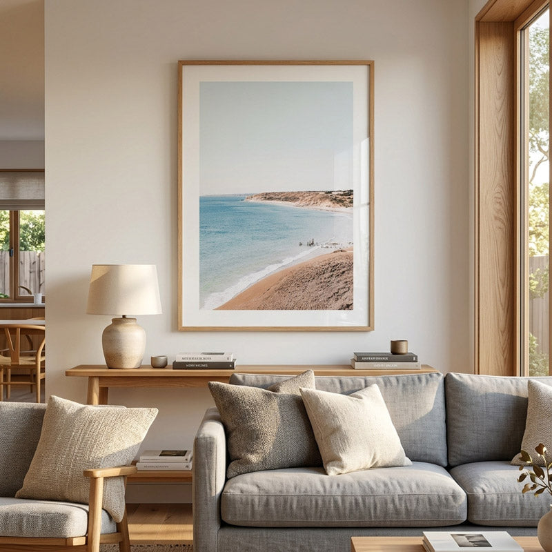 A large framed beach landscape print hangs on a beige wall above a gray sofa in a modern living room. The artwork shows a sandy beach, turquoise water, and a pale sky, with a light wood frame and white mat.