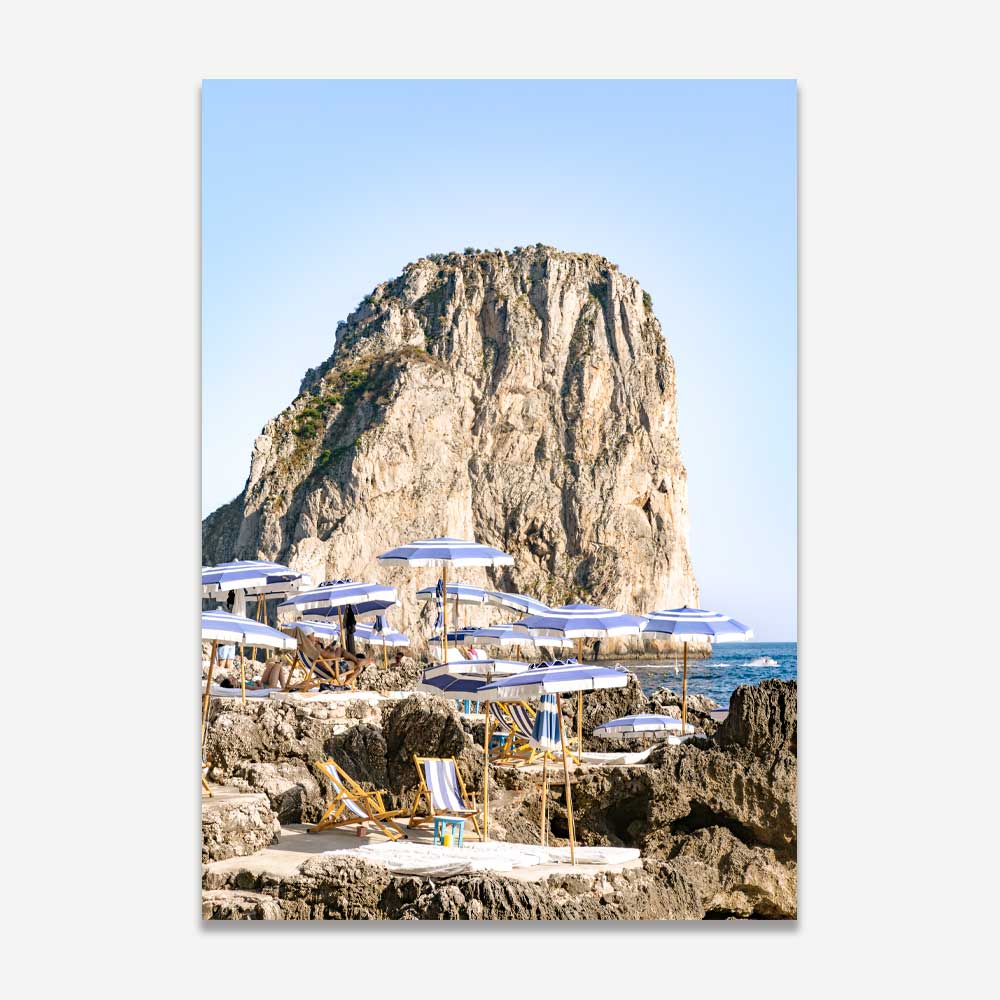 Late afternoon photograph of La Fontelina Beach Club in Capri, featuring blue and white umbrellas on rocky shores with Faraglioni rocks in the backdrop, evoking Mediterranean elegance and coastal charm.