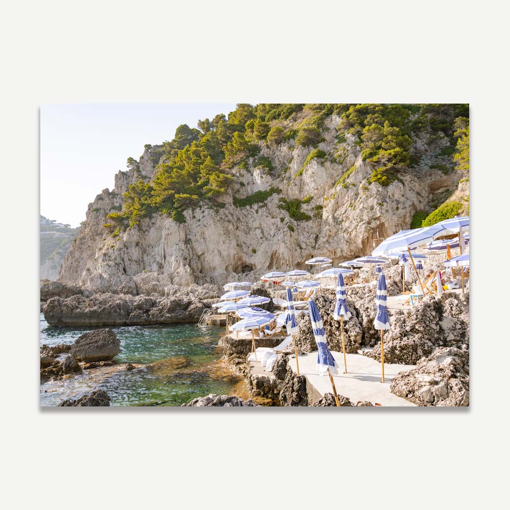 La Fontelina Beach Club in Capri with blue and white beach umbrellas and sun desks on a rocky Mediterranean shore, bathed in a golden late afternoon light, evoking coastal elegance and tranquil Italian summer vibes.