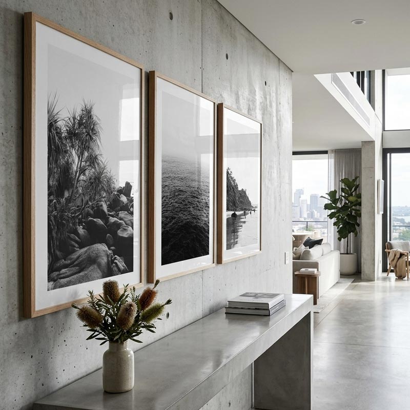 Three framed black and white landscape photographs with light wood frames are displayed on a concrete wall in a modern, minimalist living space.