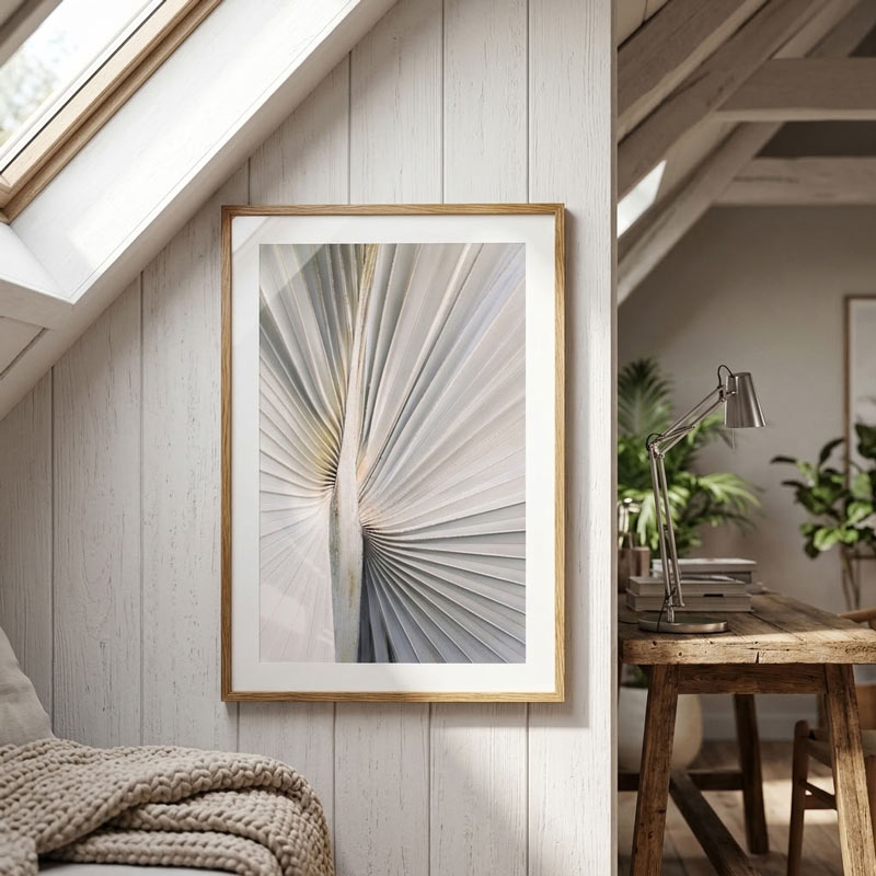 A framed abstract botanical art print featuring a sunburst pattern in soft gray, white, and beige tones, styled on a white wood paneled wall in a bright attic room with a wooden desk and plants.