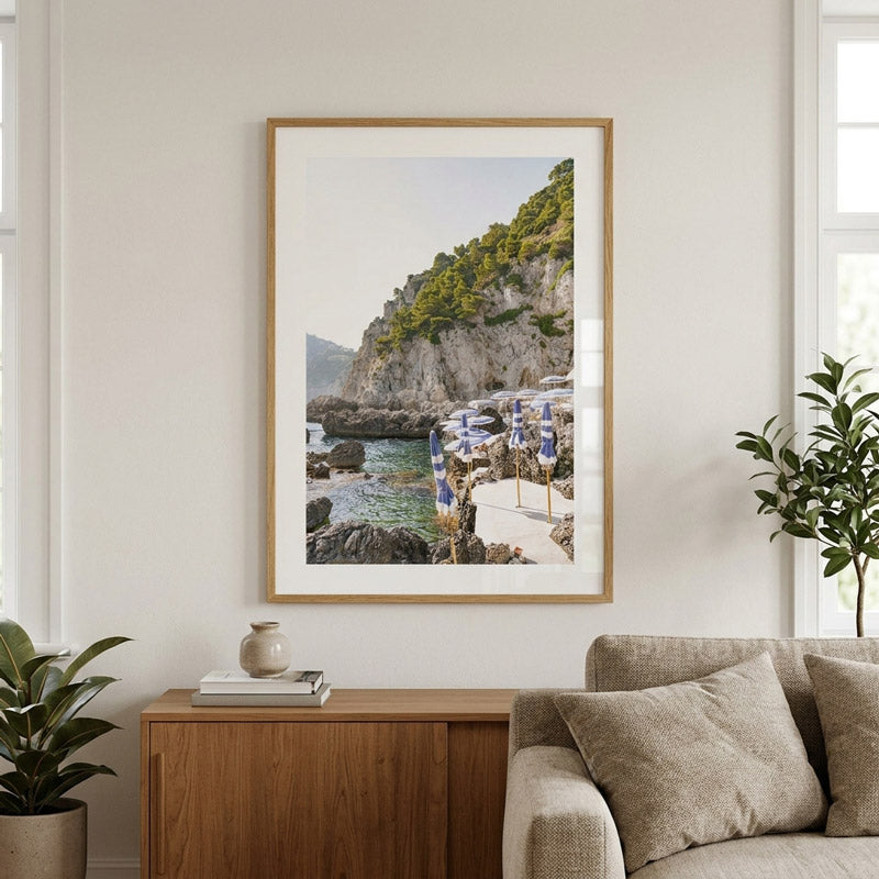 A framed coastal art print hangs on a light beige wall above a wooden sideboard, with a sofa and potted plants in the room. The print shows a rocky seaside cliff, blue water, and beach umbrellas.