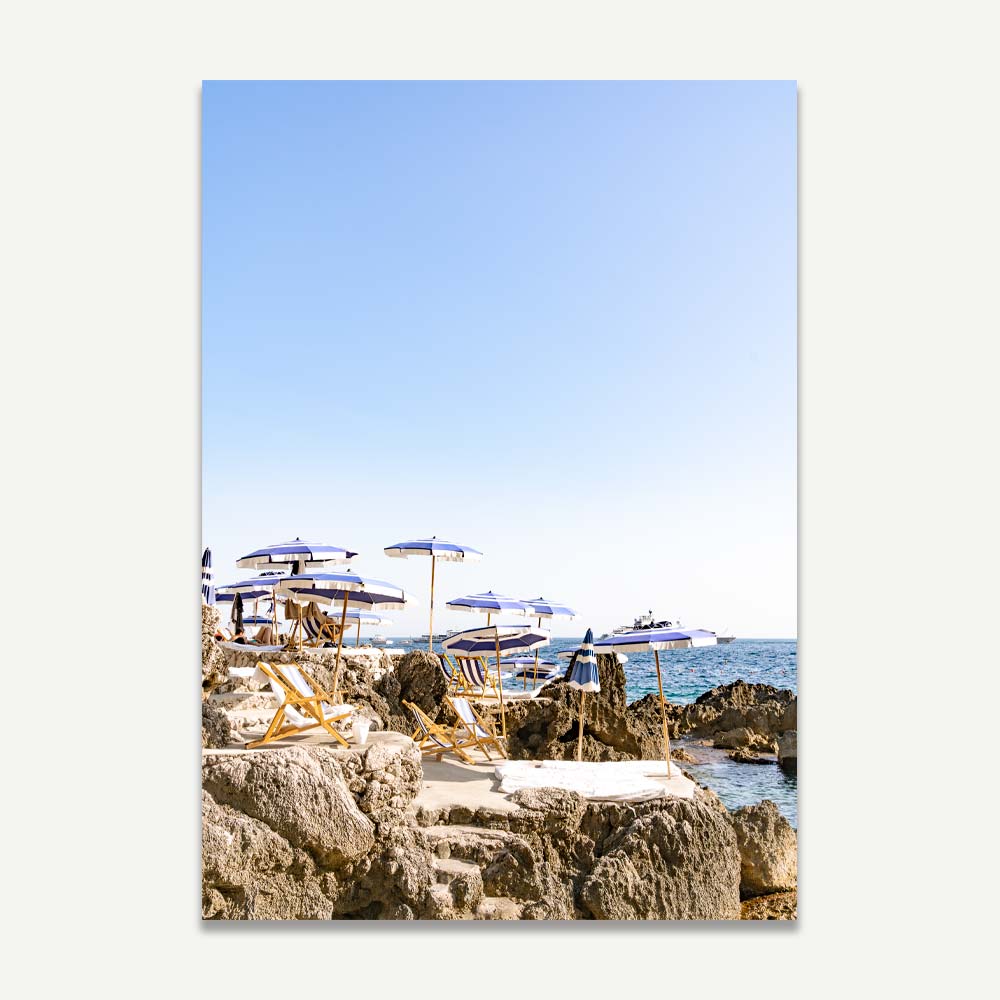 La Fontelina Beach Club in Capri with blue and white umbrellas and sun decks against the backdrop of Faraglioni rocks during a serene summer evening, bathed in warm late afternoon sunlight, evoking coastal elegance.