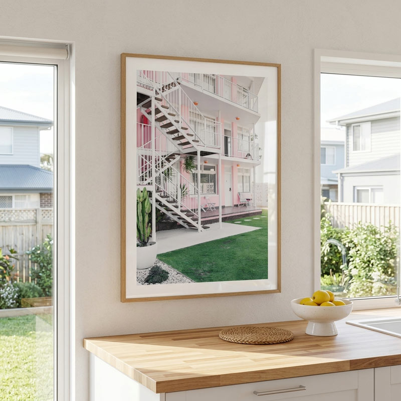 A framed art print depicting a pink apartment building with white stairs and green grass, displayed on a light beige wall above a wooden kitchen counter.