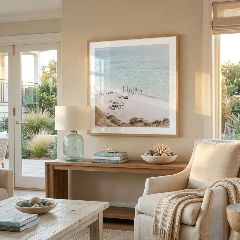 A framed coastal beach print hangs on a beige wall above a wooden console table in a styled living room with a cream armchair, glass lamp, and coastal decor.