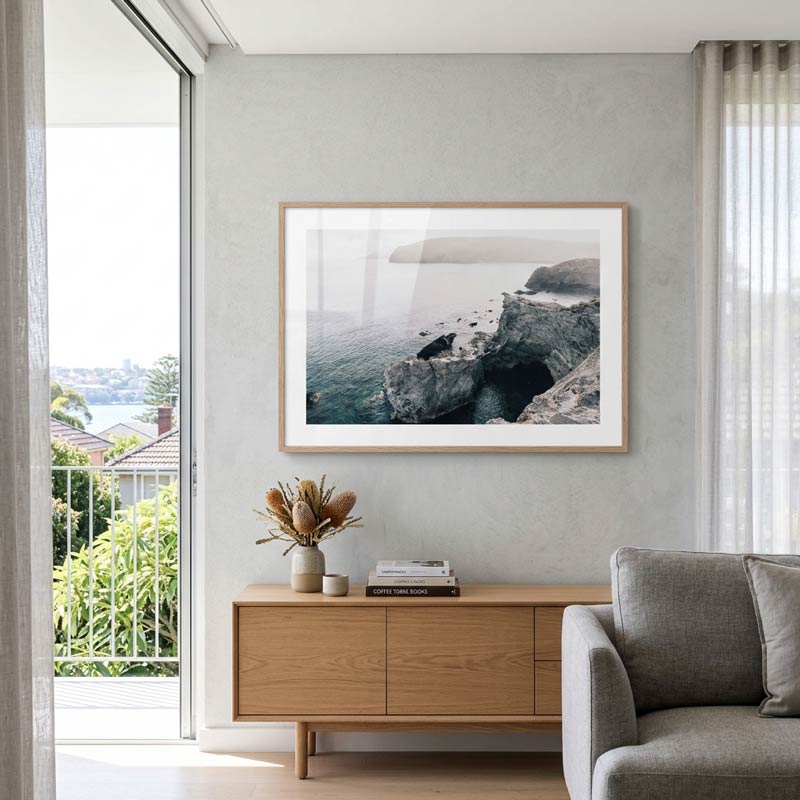A framed coastal landscape photograph hangs on a light gray wall above a wooden sideboard in a modern living room setting.