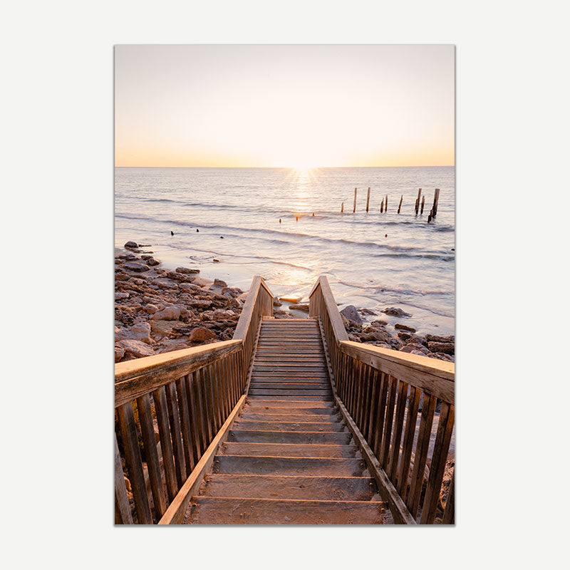Wooden staircase leading down to a rocky beach with ocean waves and sunset.