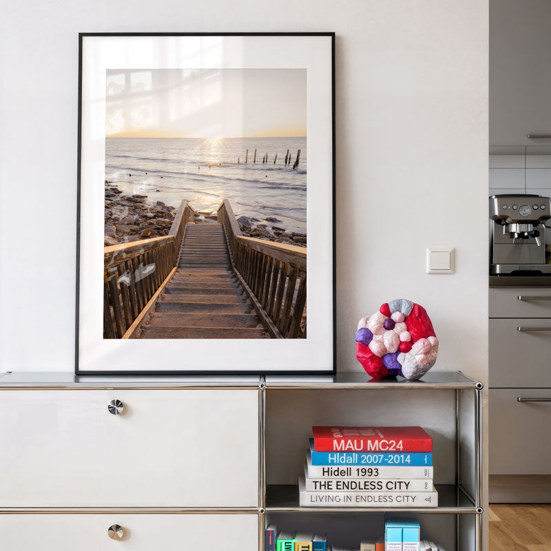 Framed photograph of a wooden pier leading to the ocean on a wall above a shelf with books and decorative items.